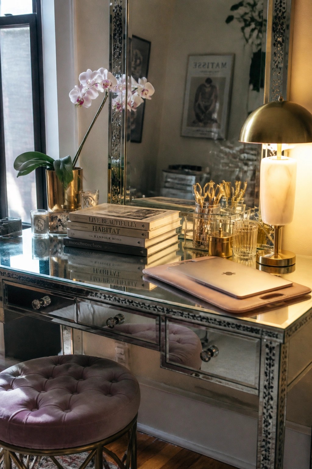 A mirrored vanity desk holding a laptop, stack of books, brass lamp, and orchid, with a purple velvet stool in front.
