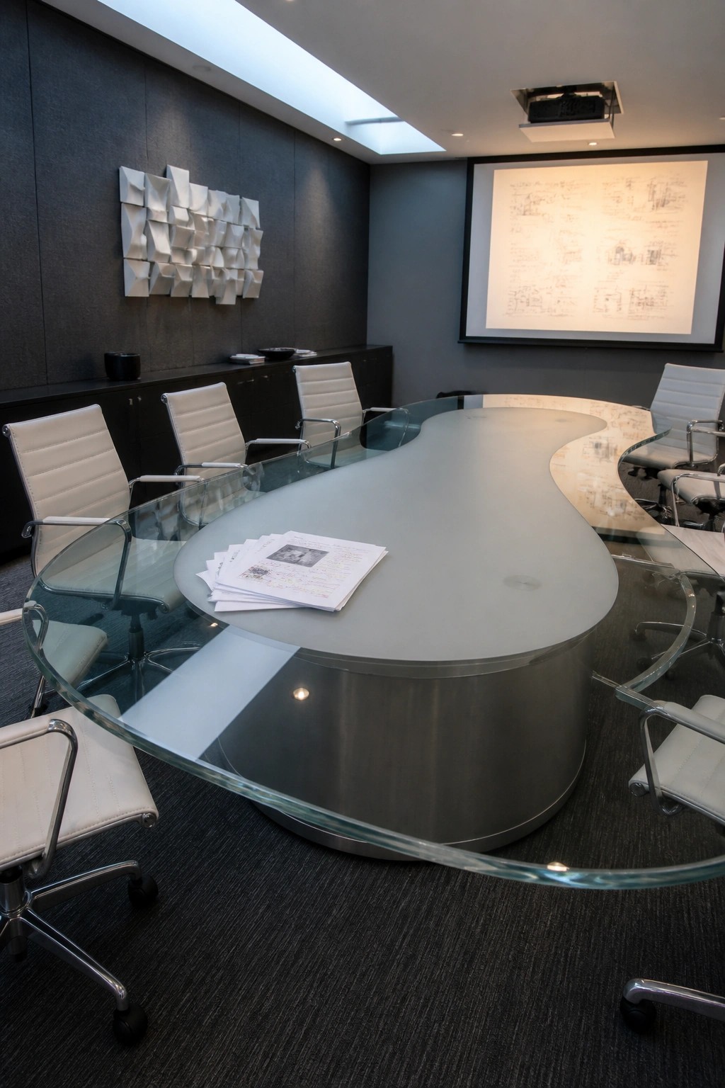 A modern conference room with a large curved frosted glass table, white leather chairs, a dark sideboard, wall art, and a projector screen showing architectural drawings.