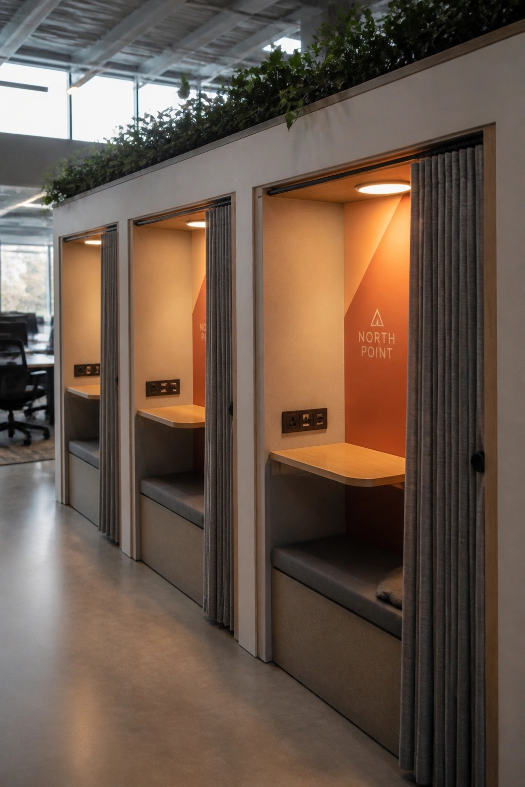 A row of modern office privacy pods with gray curtains, wooden fold-down desks, cushioned benches, warm interior lighting, and one pod showing North Point branding.