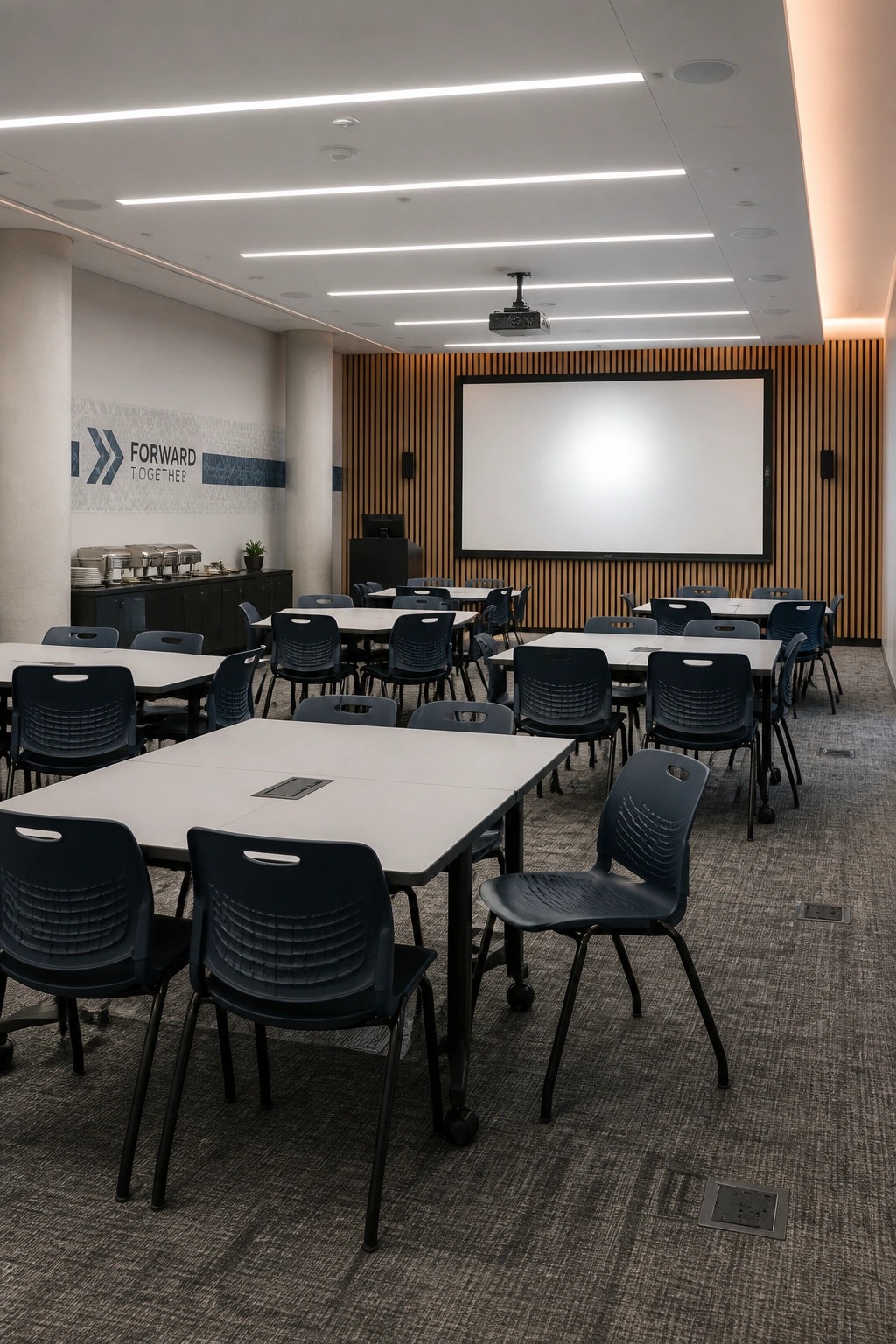 A corporate meeting room with white rectangular tables on wheels, dark blue plastic chairs, a large projection screen on a wood-slatted wall, and linear ceiling lights.