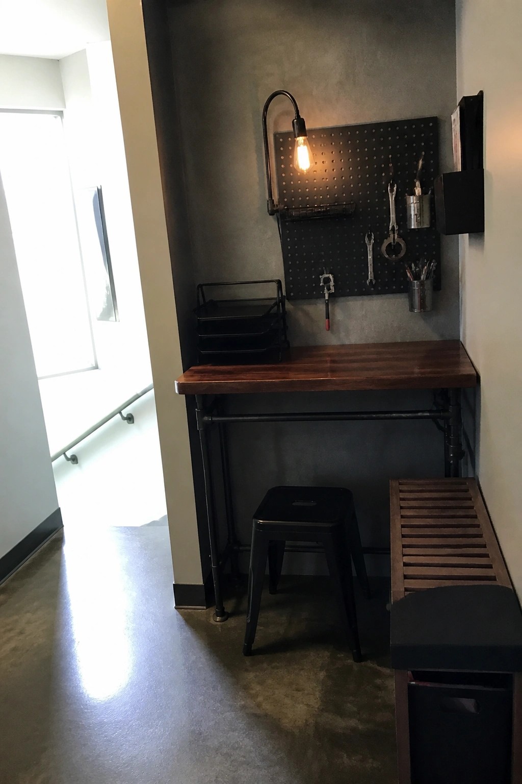 A narrow indoor workspace with a wooden desk, black pegboard holding tools on a gray wall, a black stool, and a wall-mounted light.