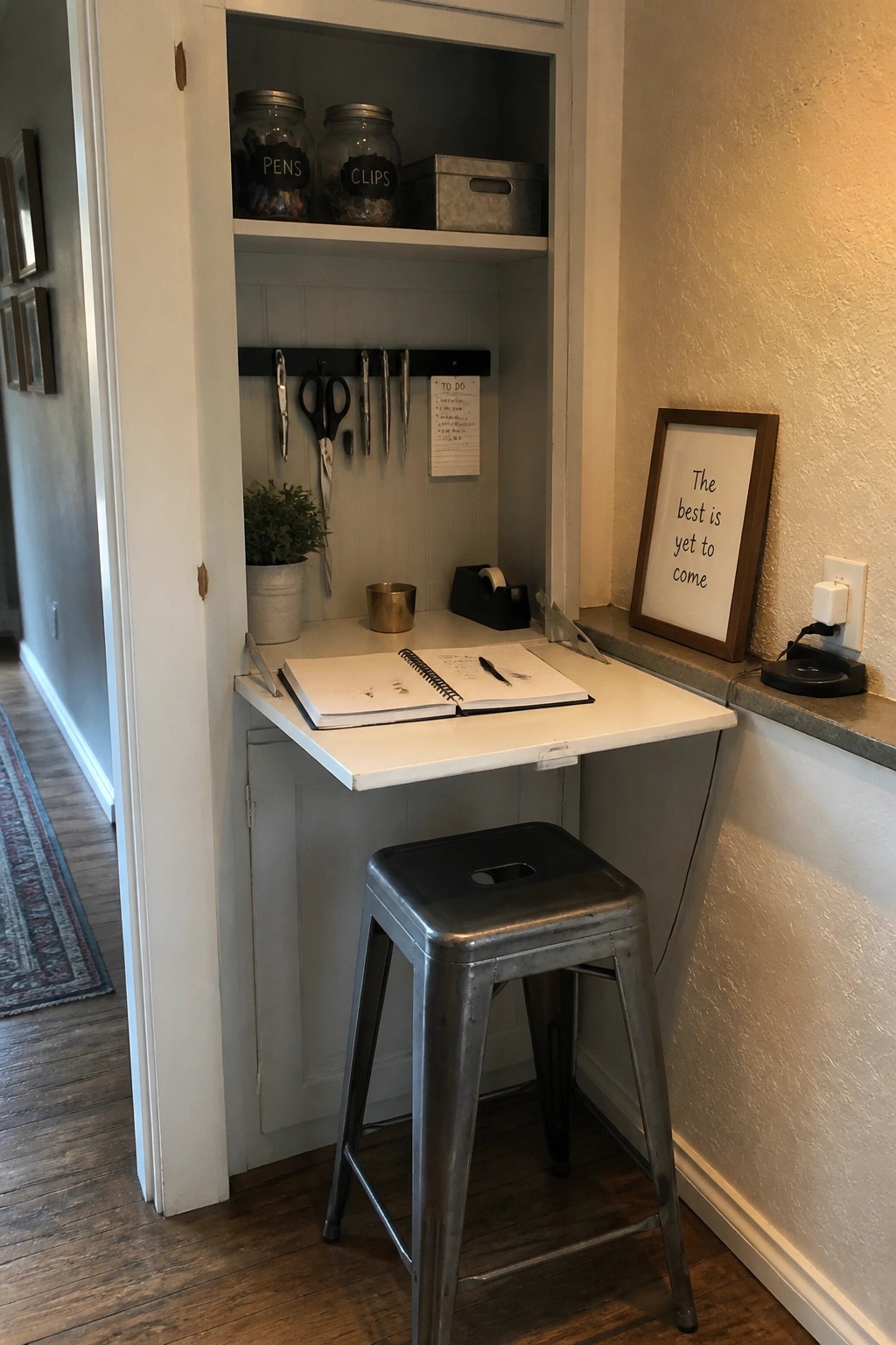 A small white fold-down desk built into a recessed gray wall nook with a metal stool underneath, office supplies on the wall, and a notebook open on the desk.
