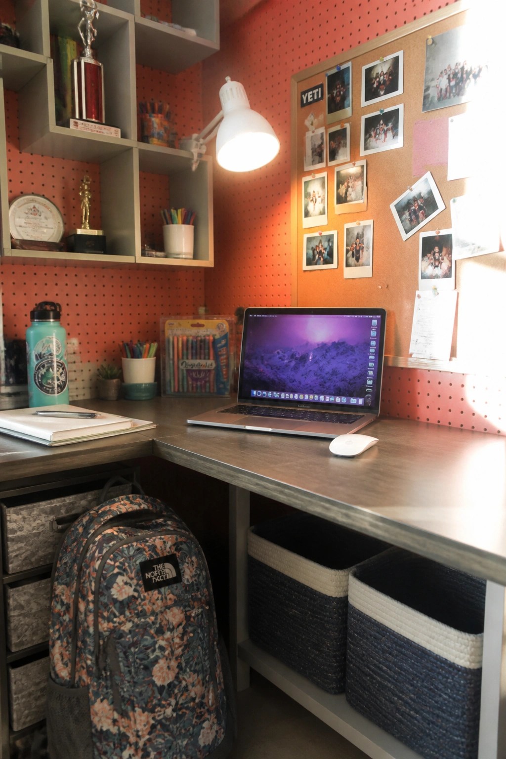 A laptop on an L-shaped desk in a corner workspace with wall shelves, a pegboard, and storage baskets below.