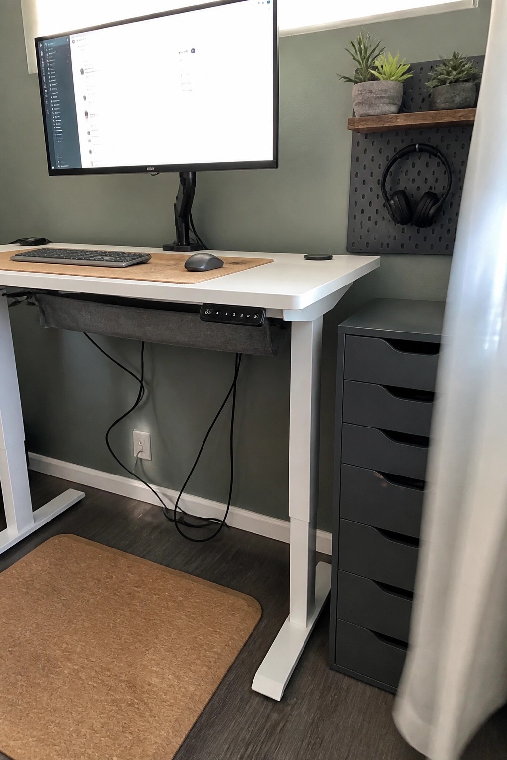 A white standing desk with monitor, keyboard and mouse sits against a green wall next to a gray multi-drawer cabinet and a black pegboard holding potted plants and headphones.