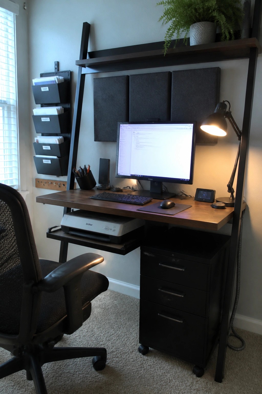A home office with a wooden desk, computer monitor, overhead black shelving unit, acoustic panels, desk lamp, and black filing cabinet on wheels.
