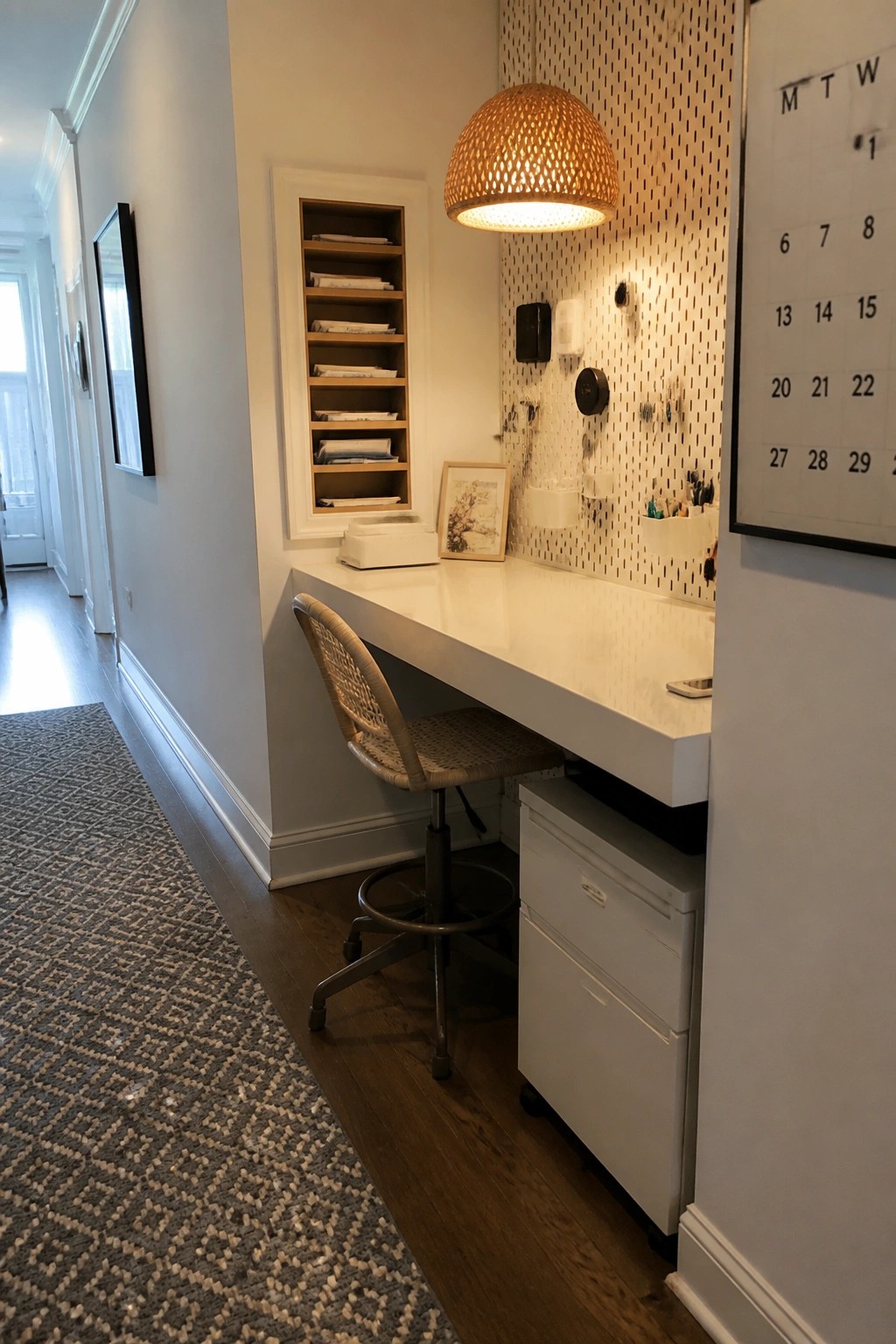 A narrow home office features a white built-in desk under recessed wooden shelves, a woven chair on wheels, a pegboard wall with small organizers, and a white filing cabinet below the desk.