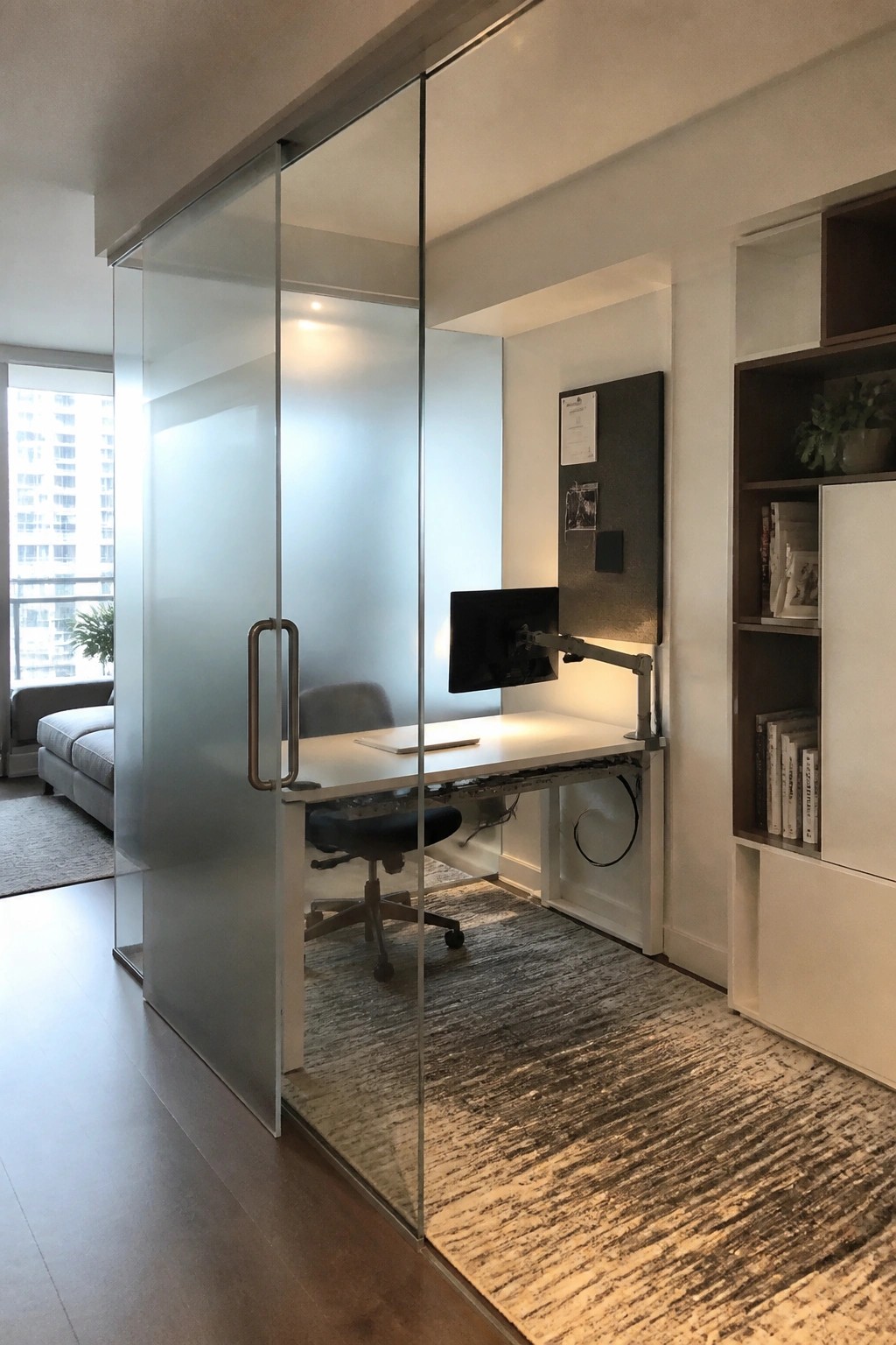 A home office space enclosed by frosted glass sliding doors, with a white desk, monitor, and office chair visible inside.