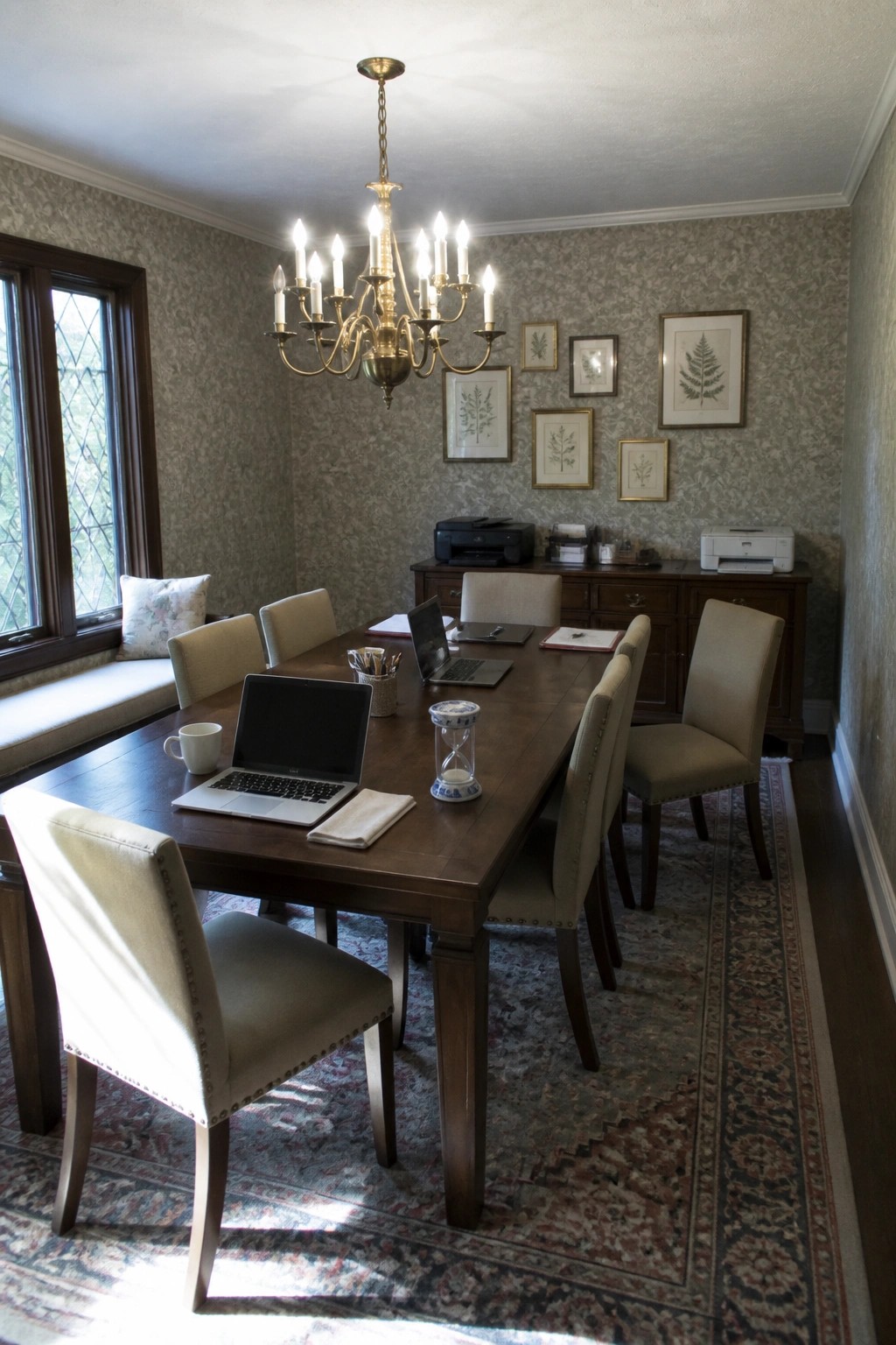 A long wooden dining table with laptops, a mug, and an hourglass, surrounded by upholstered chairs in a room with a chandelier and framed botanical prints.