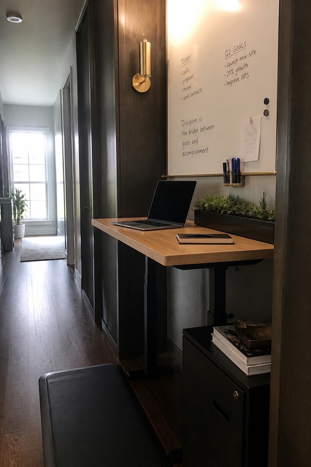 A narrow home workspace with a light wood desk, open laptop, black cabinet below, and a large whiteboard mounted on the wall above showing handwritten notes and goals.
