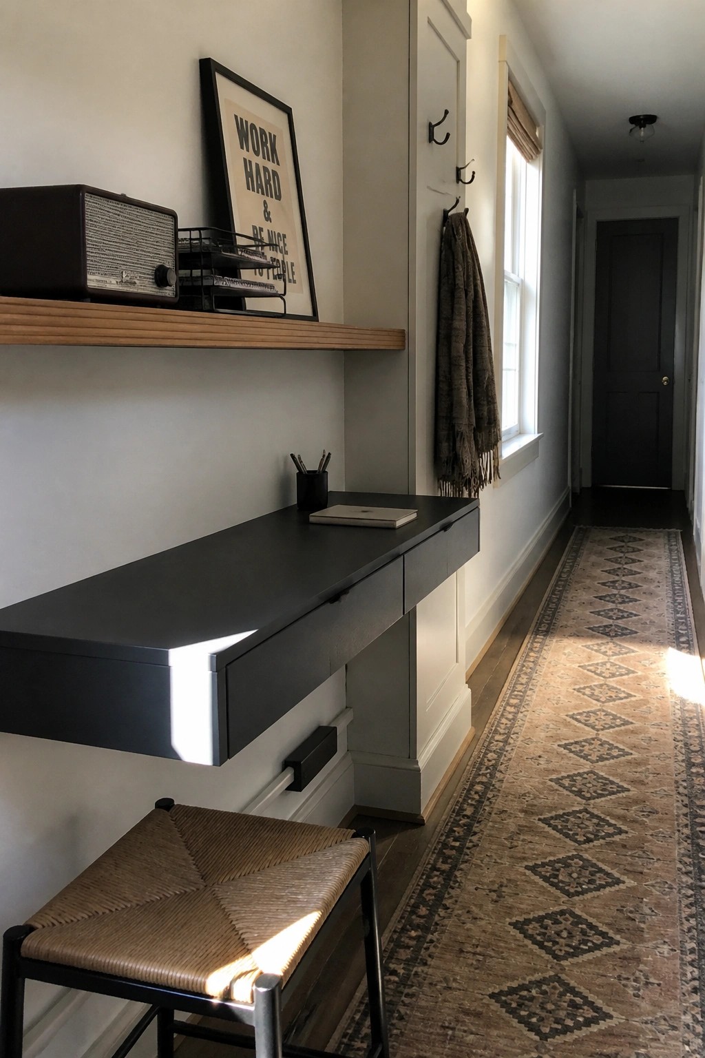 A black floating desk is mounted on a light-colored wall in a narrow hallway, with a woven stool tucked underneath, a wooden shelf above holding a radio and framed print, and a long patterned runner on the floor.