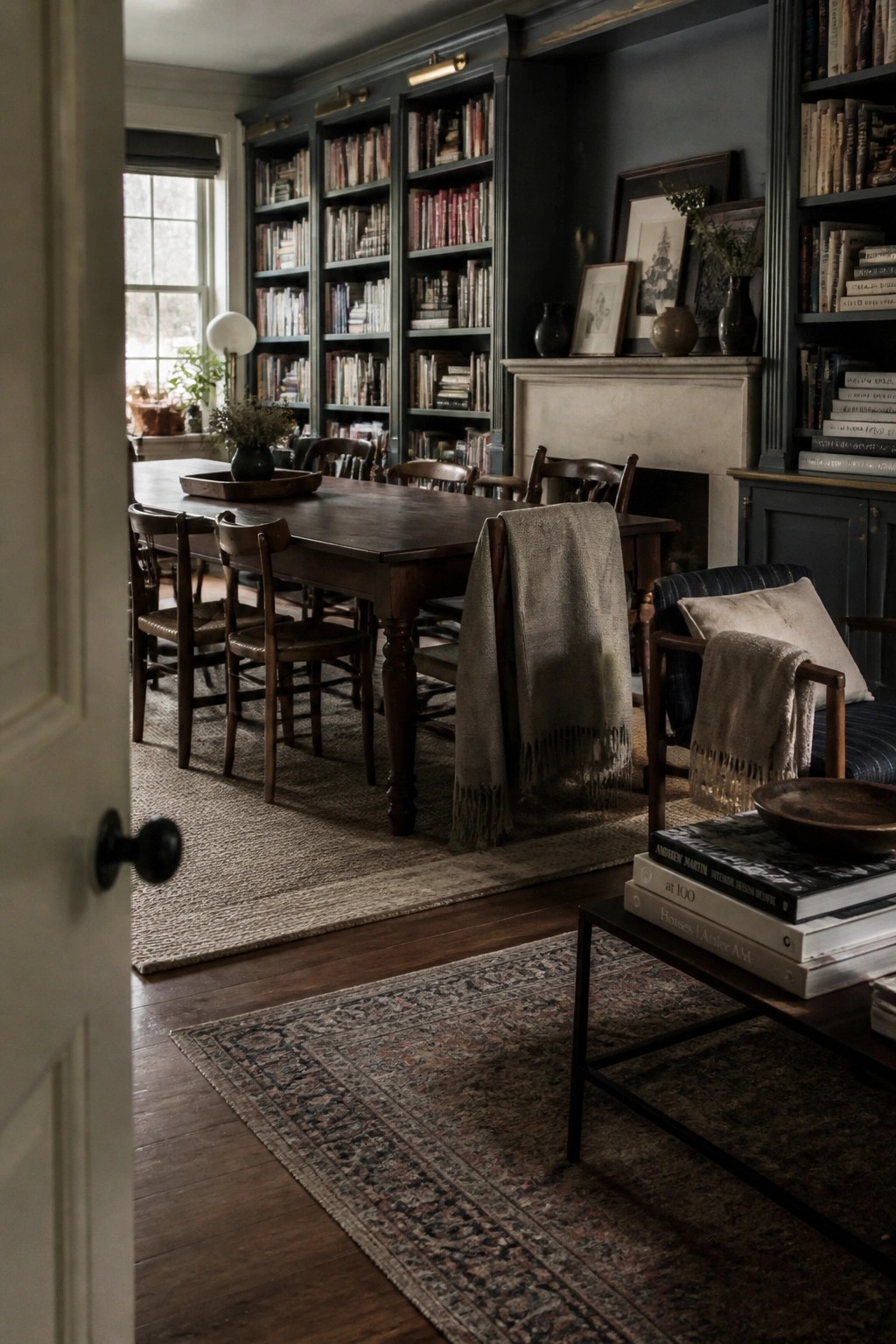 A library room with dark built-in bookshelves filled with books, a long wooden table and chairs on layered rugs, and a fireplace with framed art on the mantel.