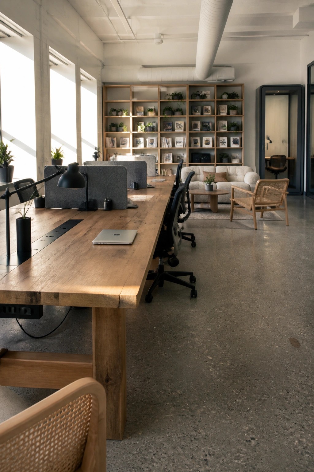 A modern office interior with a long wooden communal table, black office chairs, a laptop, desk lamps, and a background wall of wooden shelves filled with books and plants plus a white sofa area.