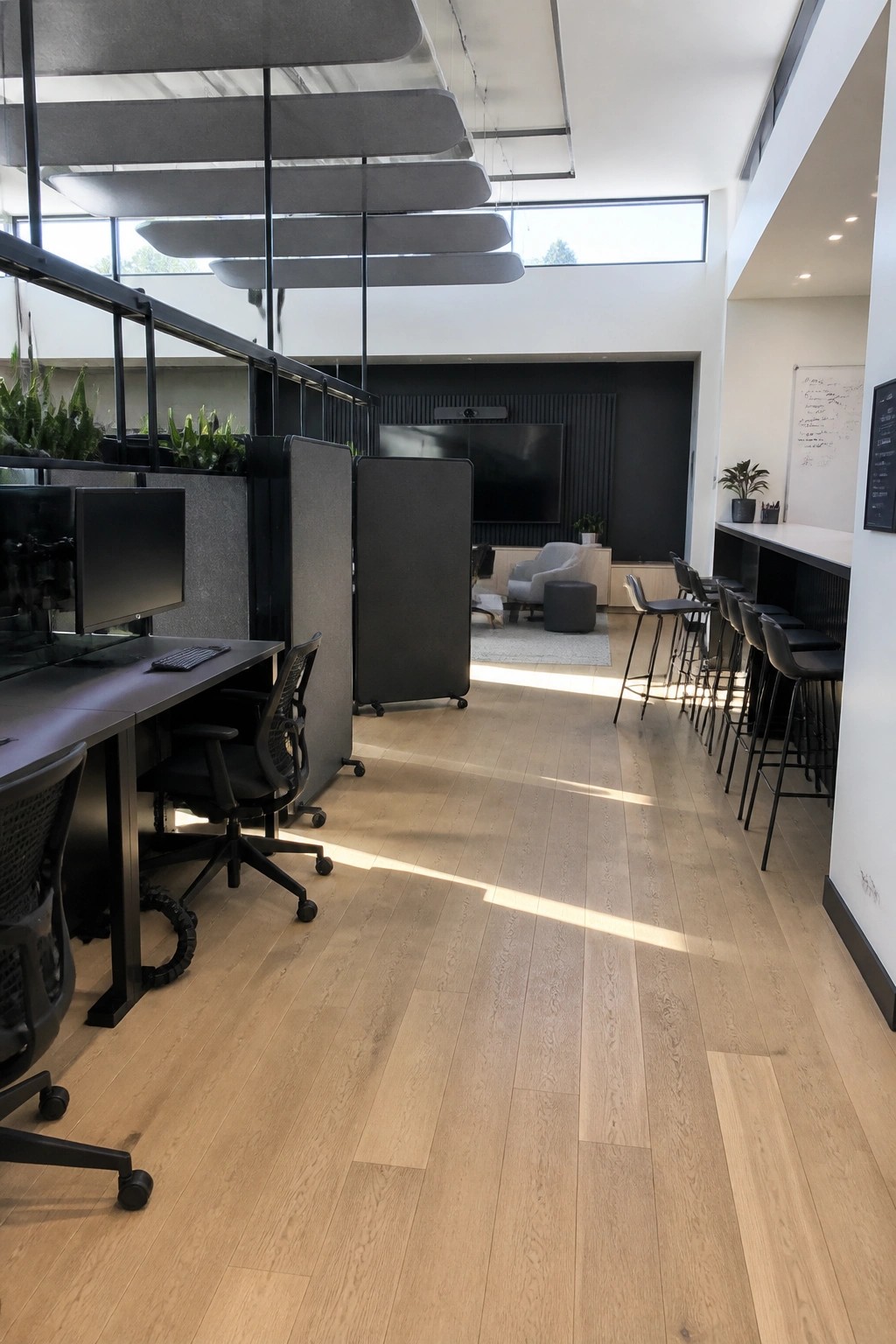 Modern open-plan office with individual desks separated by gray acoustic panels, a lounge area with seating and a television, and a long bar counter with stools along the right wall.