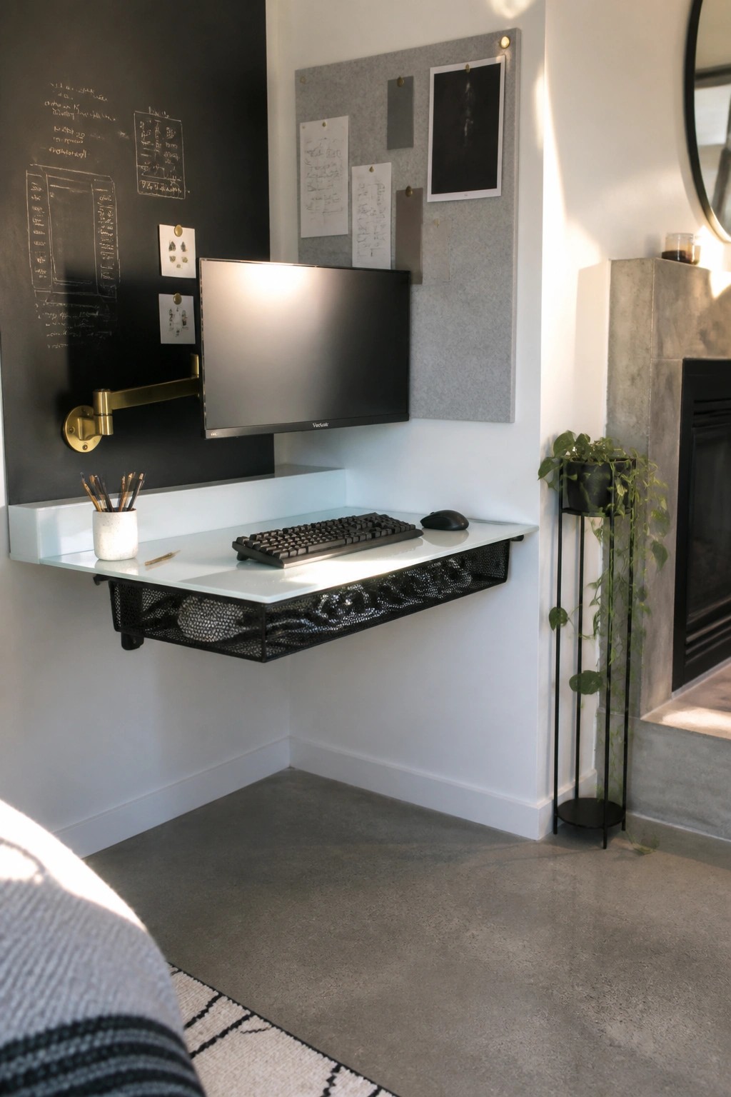 A compact wall-mounted desk with a monitor on an arm, keyboard, and under-shelf storage in a corner with a chalkboard wall nearby.