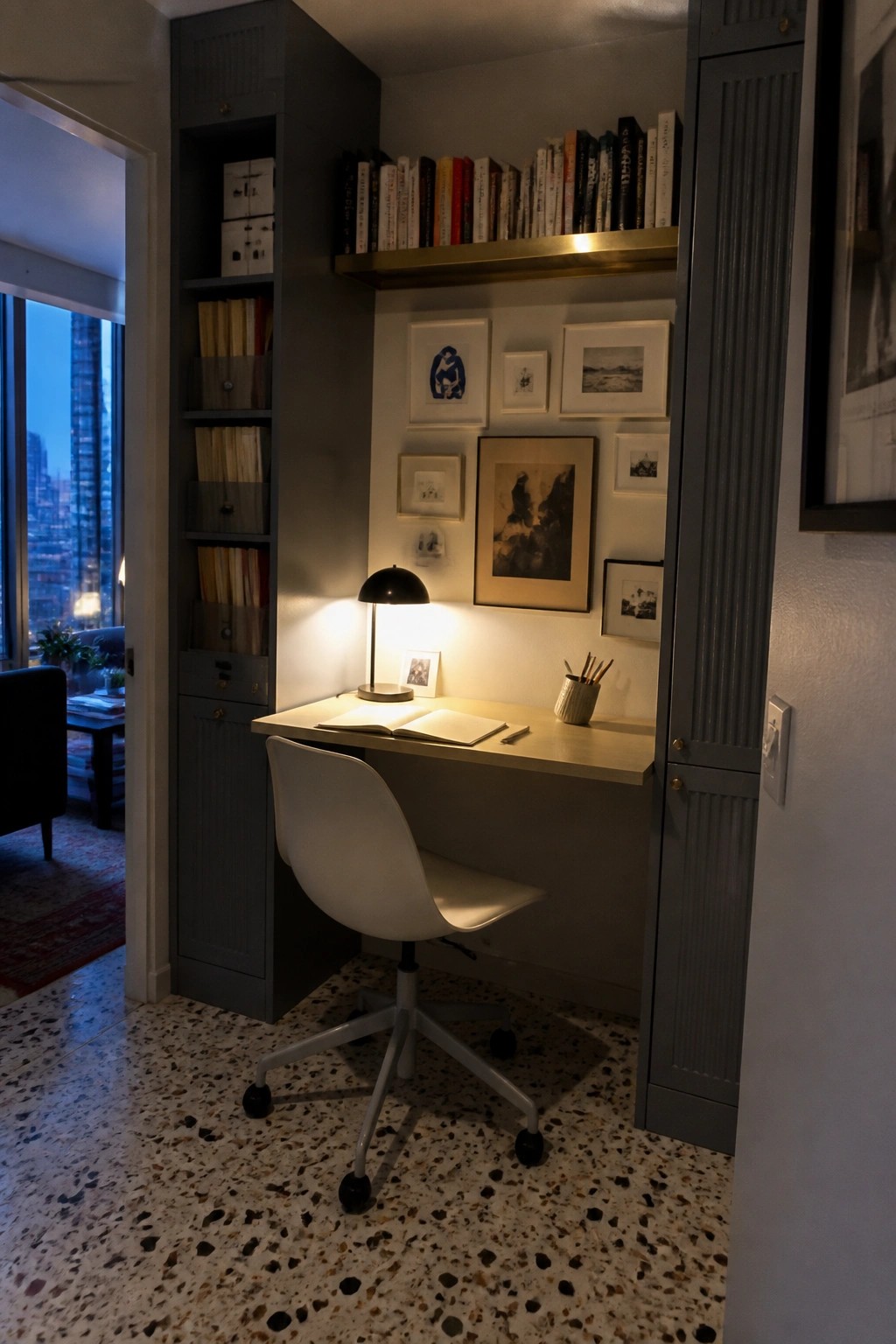 Built-in desk nook with white chair, lit lamp, open notebook, bookshelves, and framed art.