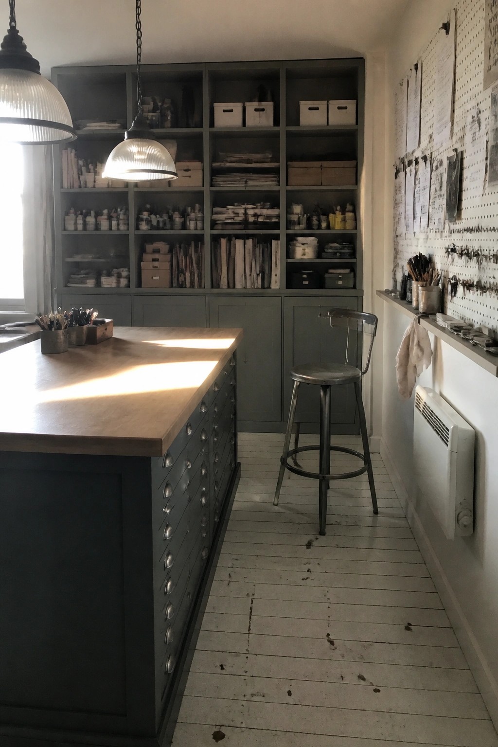 A narrow home office with a large wooden work table, gray floor-to-ceiling built-in shelves and cabinets, a metal stool, and a pegboard wall holding papers and tools.