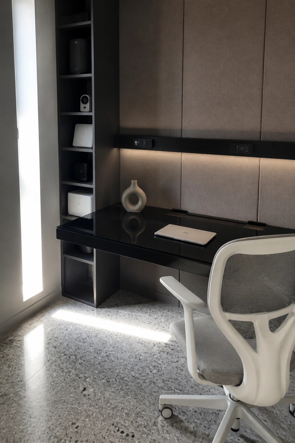 A modern workspace with a built-in black desk, integrated shelving, upholstered wall panels, and a white mesh office chair on a speckled floor.