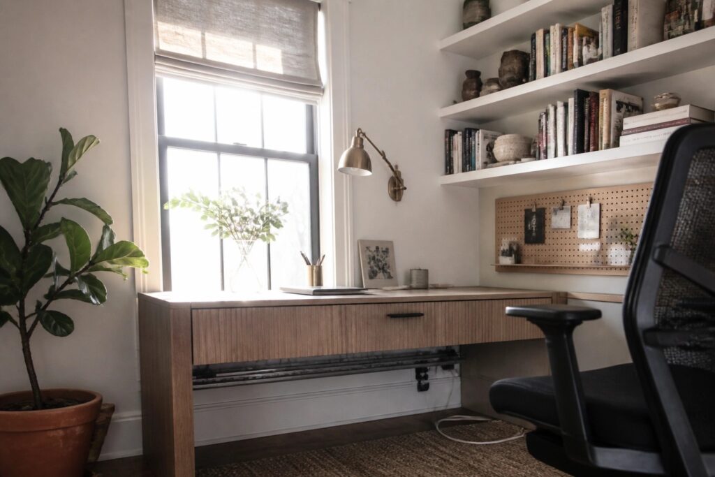 17 Inspiring Workspace Ideas for Productive and Beautiful Everyday Setups A light wood desk with drawers sits under a window, topped by two white floating shelves filled with books and pottery, next to a pegboard and a potted plant.