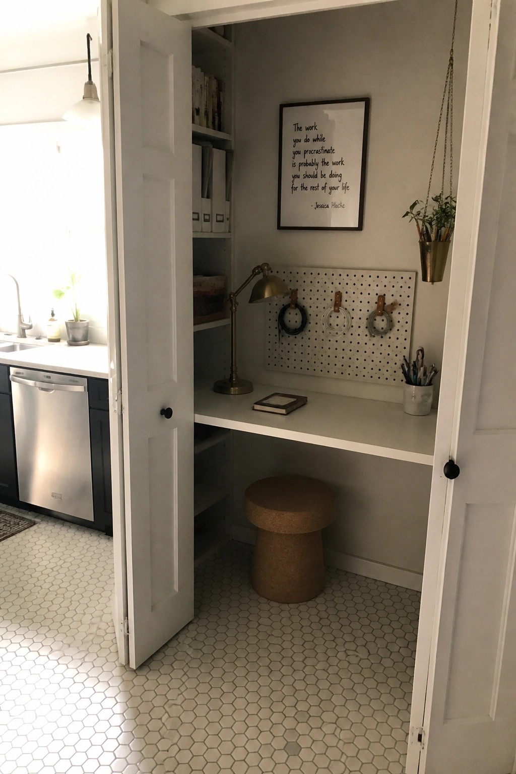 A small built-in desk inside an open closet with white doors, showing a pegboard on the wall, desk lamp, notebook, cork stool, and shelves above.