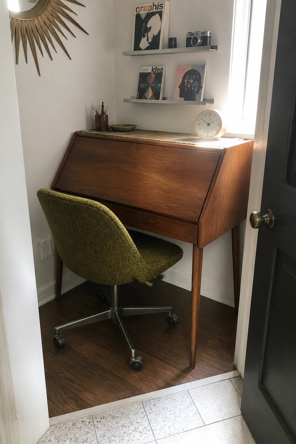 A wooden drop front desk with a green upholstered swivel chair sits in a small corner beneath two floating shelves holding books and a camera.