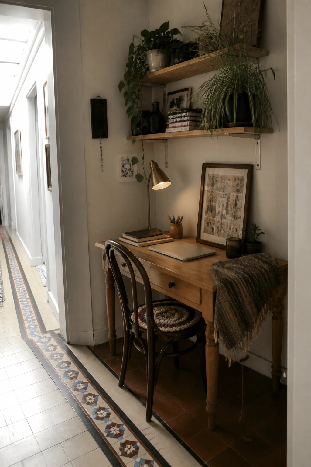 A small wooden desk and chair sit in a narrow indoor hallway corner beneath two wooden wall shelves holding plants and books, with a lit desk lamp and framed pictures nearby.