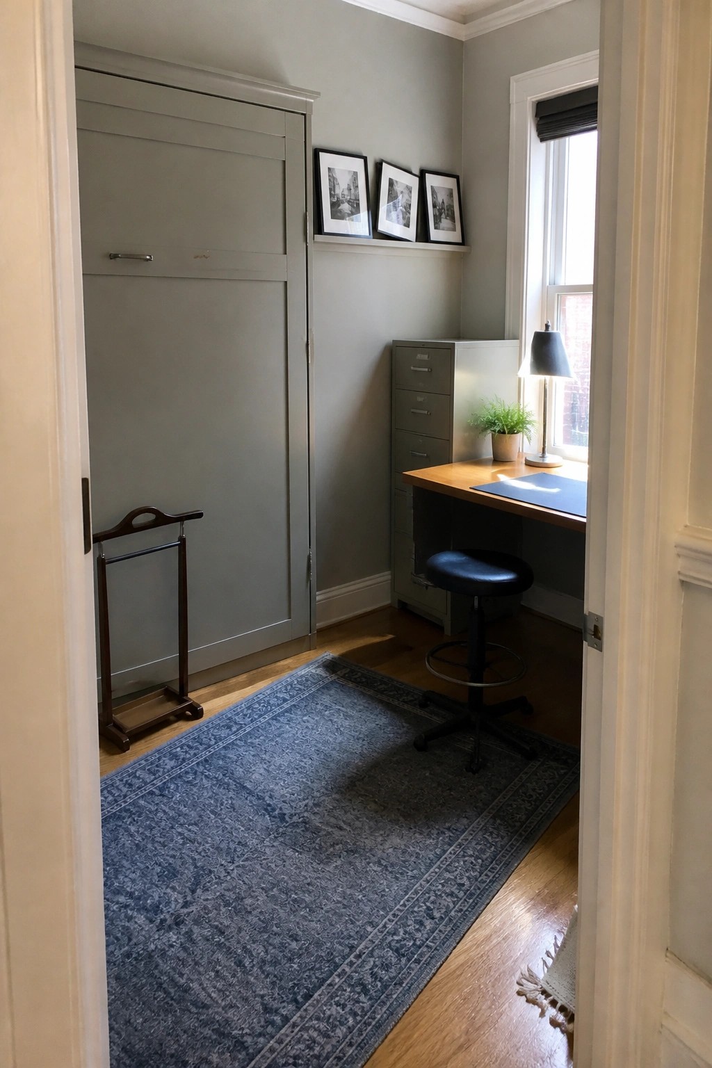 A compact room with a tall gray wardrobe on the left, a wooden desk under a window, a black stool, a gray filing cabinet, and a blue patterned rug on hardwood floors.
