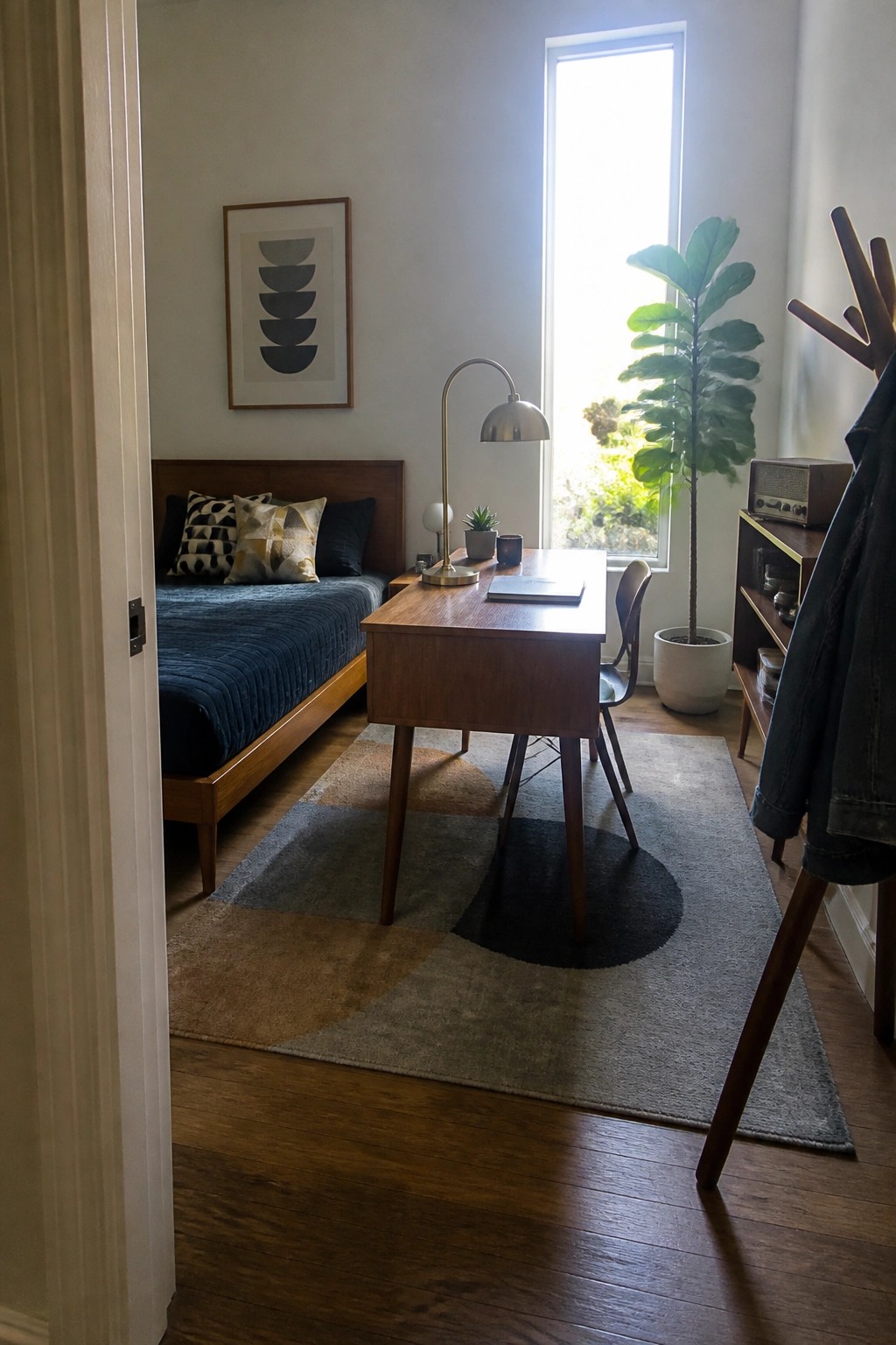 A compact bedroom interior showing a wooden desk placed on a large patterned rug, with a bed to the left and a tall window behind the desk.