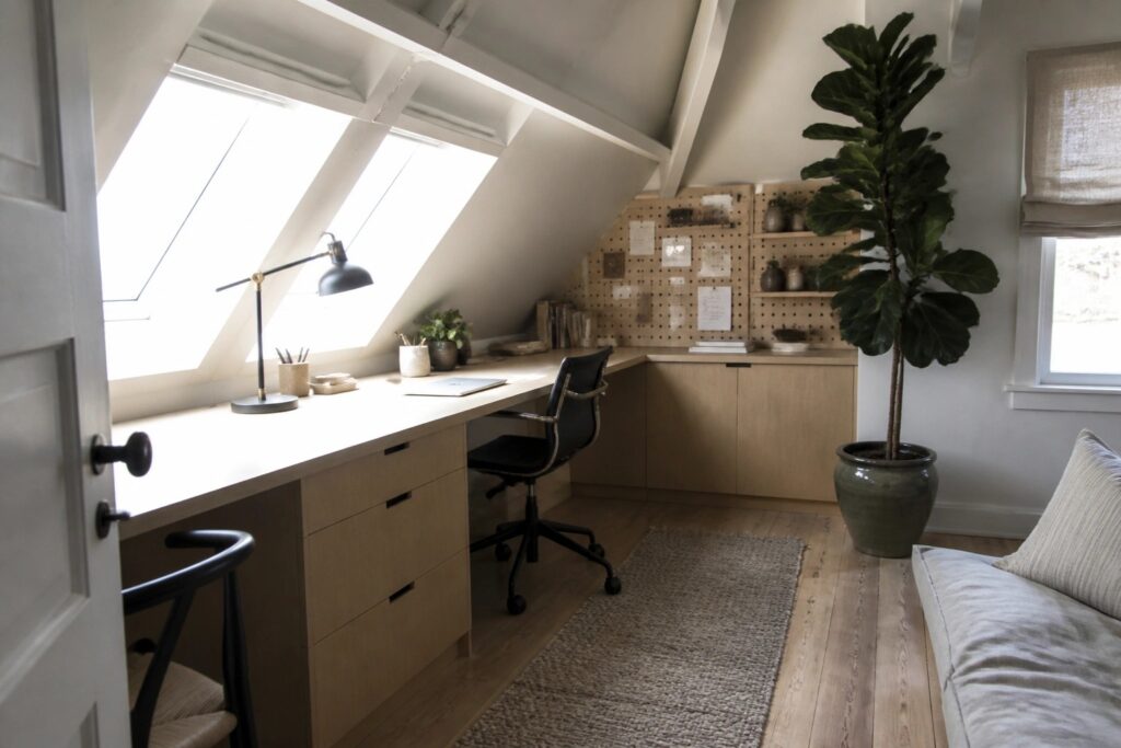 An attic workspace features a long light wood desk beneath multiple skylights, a black office chair, a pegboard on the sloped wall, and a large potted plant near a window with a rolled shade.