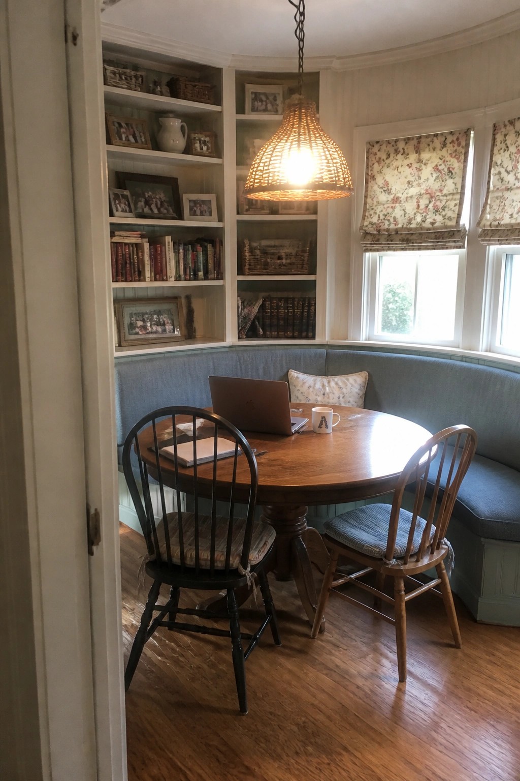 Cozy round wooden table with laptop in blue bench-lined reading nook with shelves.