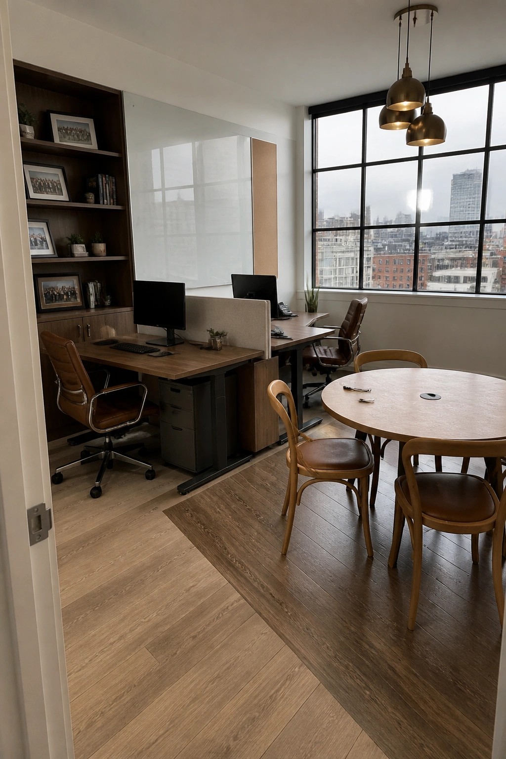 An executive office with two wooden desks and monitors, brown leather chairs, a round wooden meeting table, built-in bookshelves on the left, and large windows showing a city view.