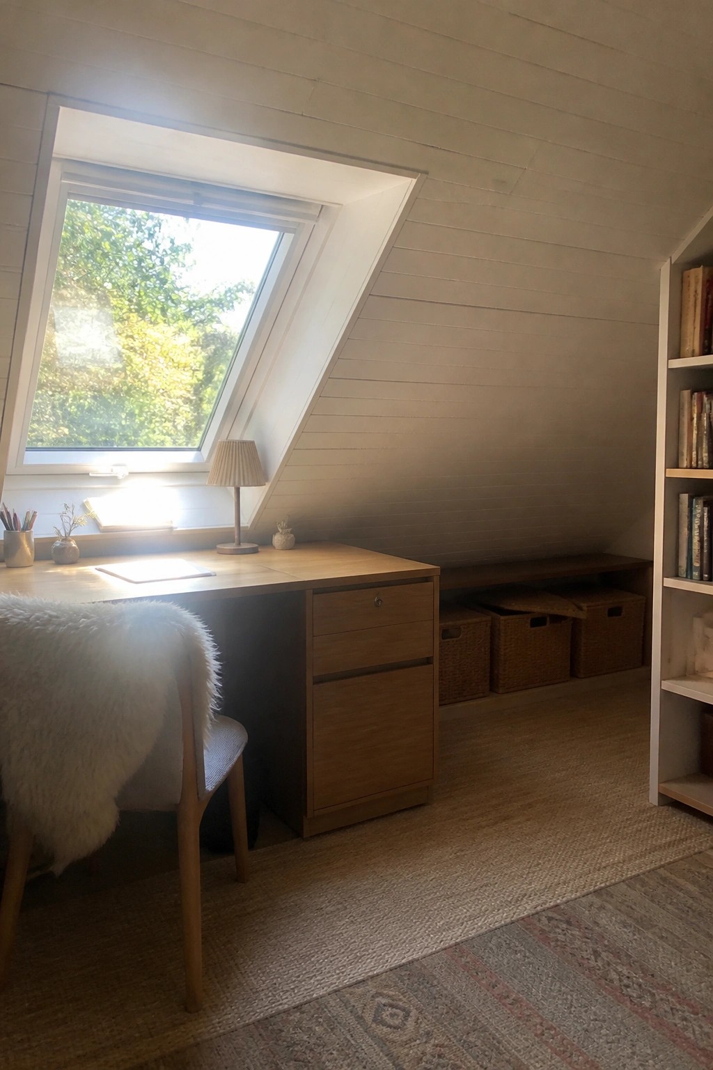 A wooden desk with drawers sits under a large slanted window in an attic room, with a chair on the left and woven baskets stored along the low wall to the right.