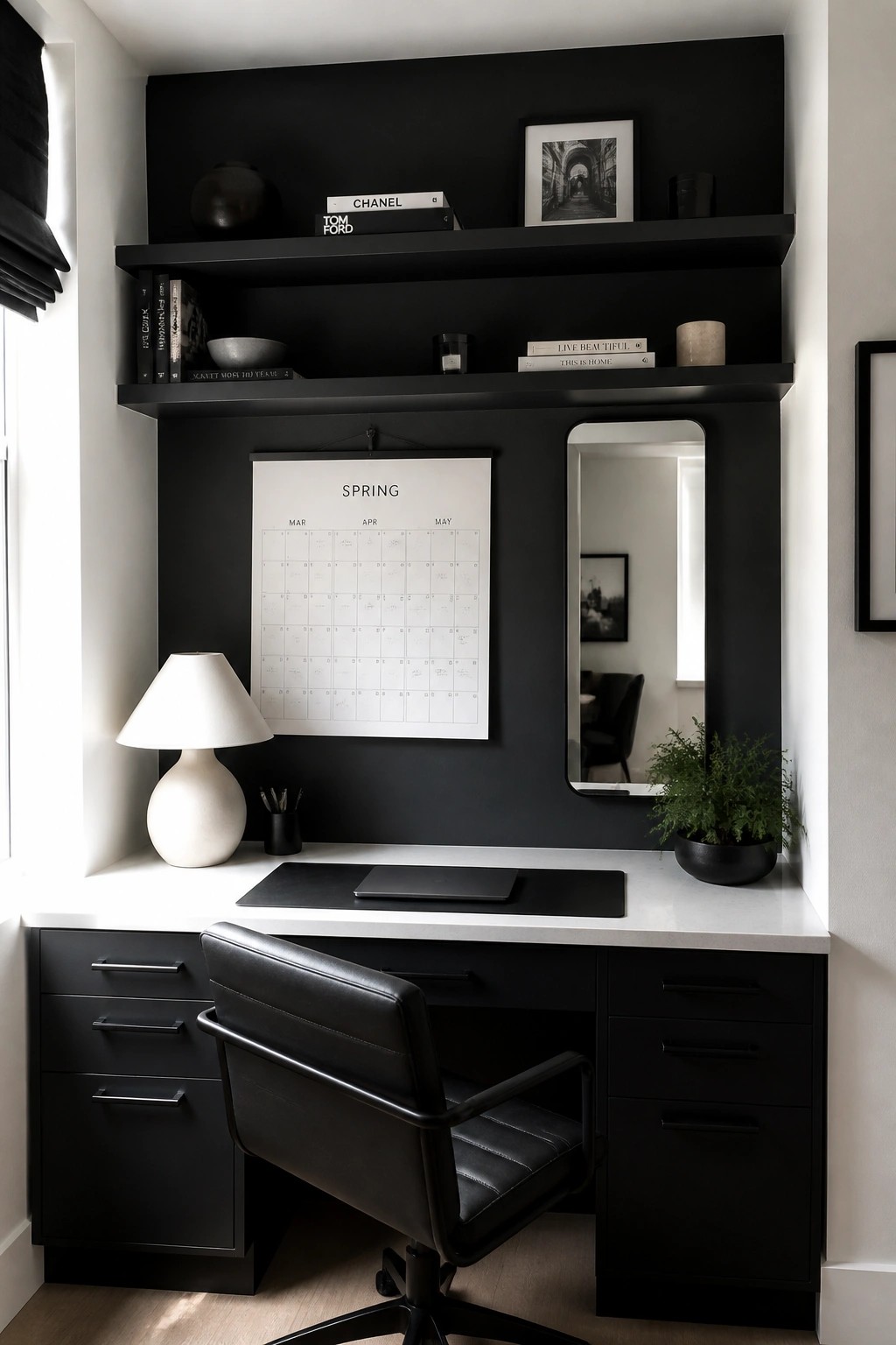A home office featuring a black built-in desk with drawers and overhead shelves against a black wall, a white spring calendar, a mirror, a white lamp, and a black office chair.
