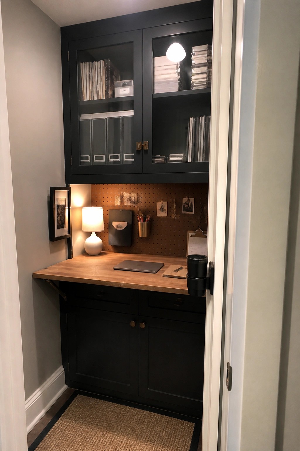 Compact home office nook with dark cabinets, wooden desk, pegboard wall, and lit lamp