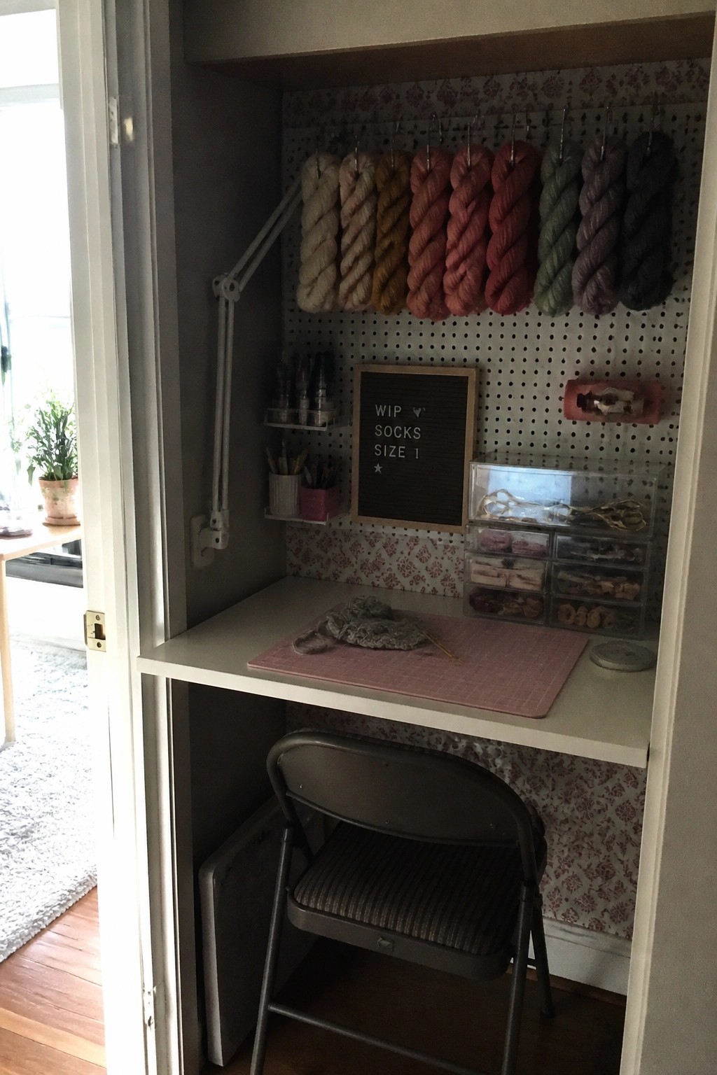 A small built-in desk inside a closet with a pegboard holding rows of yarn, craft supplies on shelves, a letter board, and a folding chair pulled up to the desk.