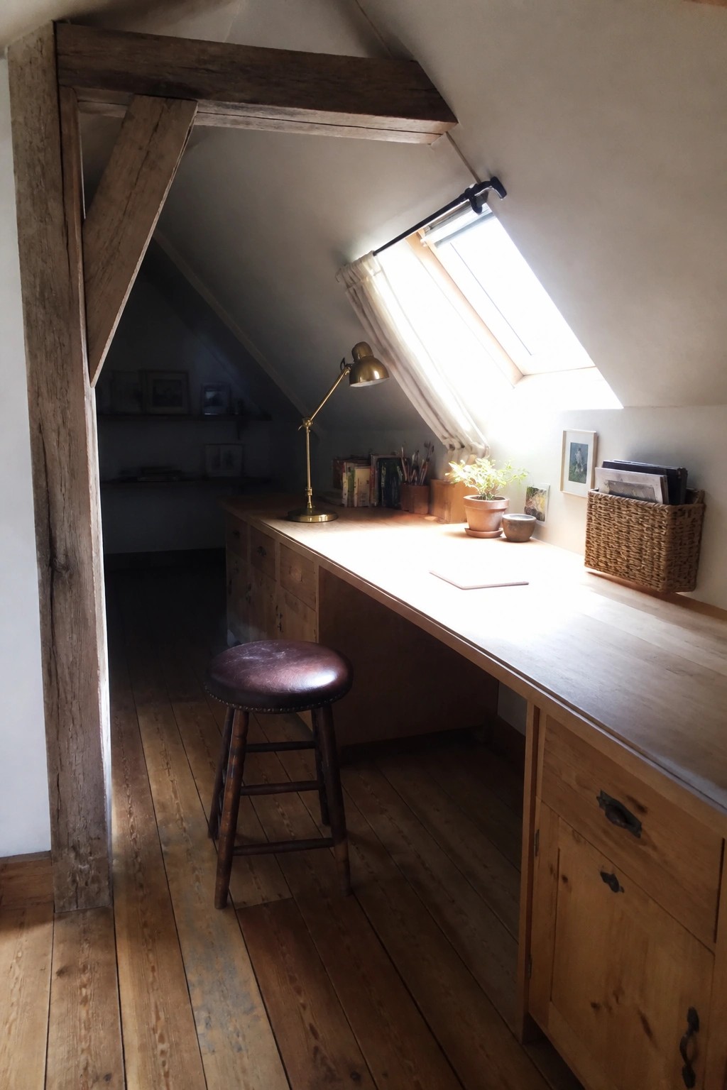 A long wooden desk runs beneath a skylight in an attic room with exposed beams, a brown leather stool underneath, and storage drawers built into the desk.