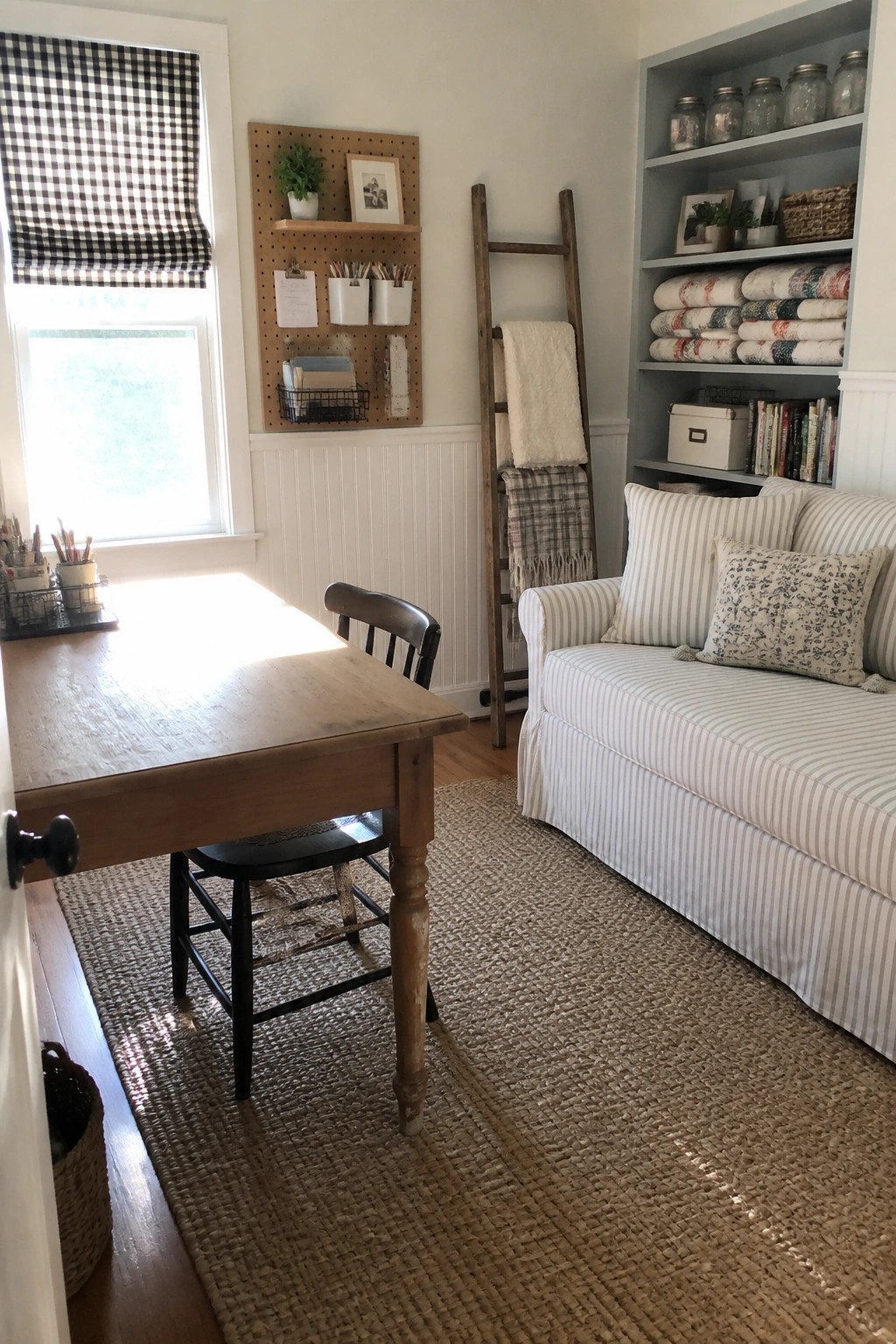 A home office interior showing a wooden desk with a black chair on a woven rug, a striped sofa against the right wall, a pegboard above wainscoting, and built-in shelves holding blankets and books.