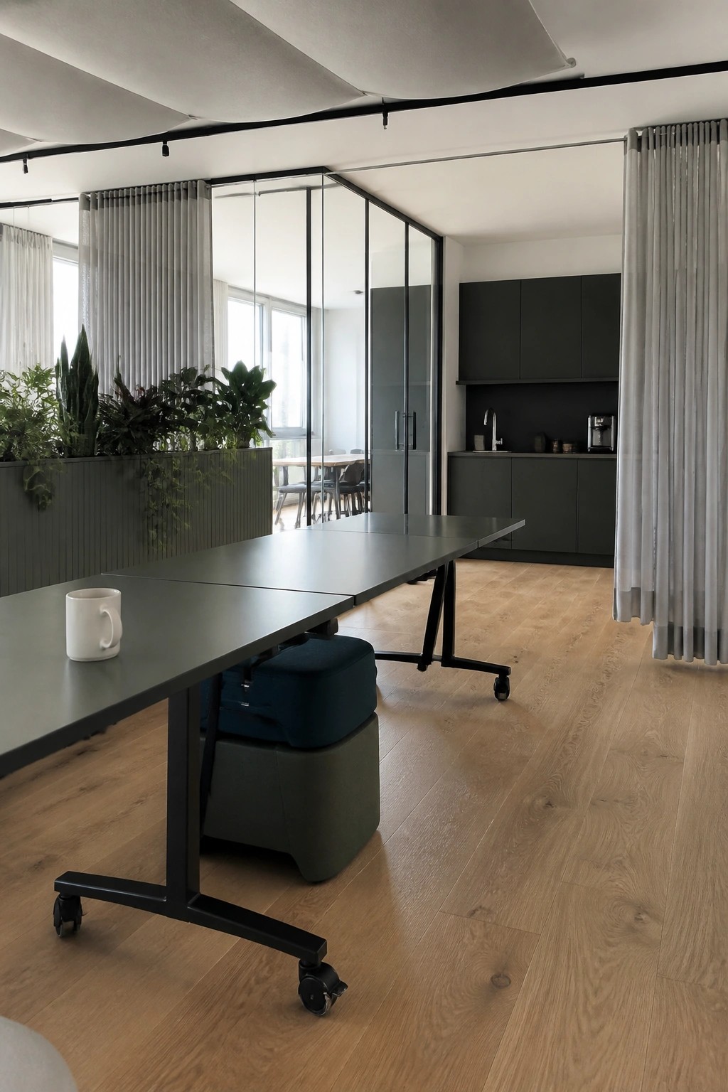 Long dark table with mug in modern office with kitchen, plants, and glass walls.