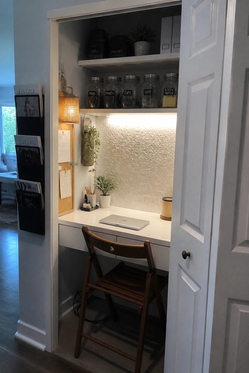 A compact white desk is built into an open closet with upper shelves holding labeled glass jars, a laptop on the desk surface, and a wooden folding chair pulled up to the desk.