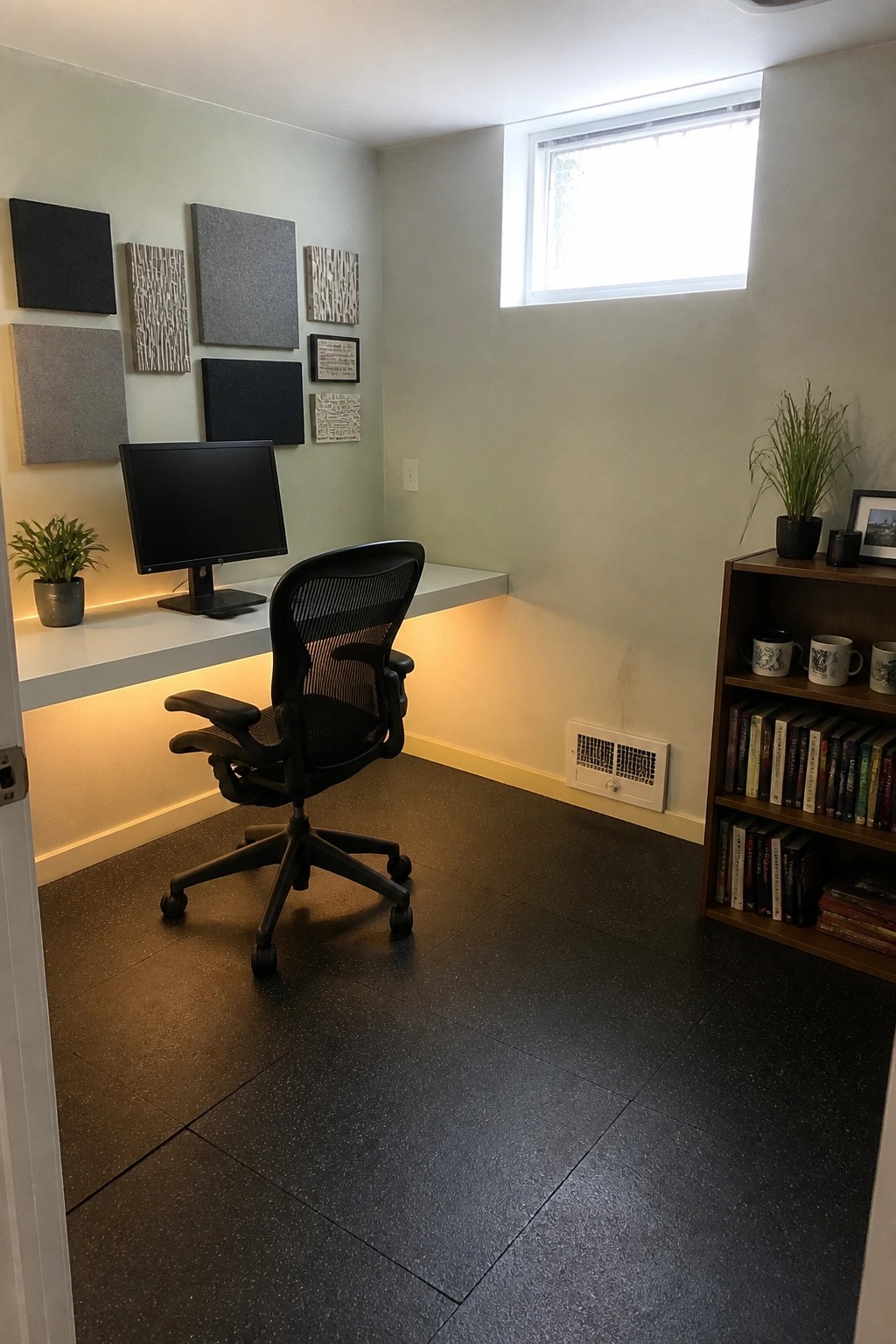 A home office with a white floating desk lit by under-shelf lighting, a black ergonomic chair, gray acoustic panels on the wall, and a window above the desk area.