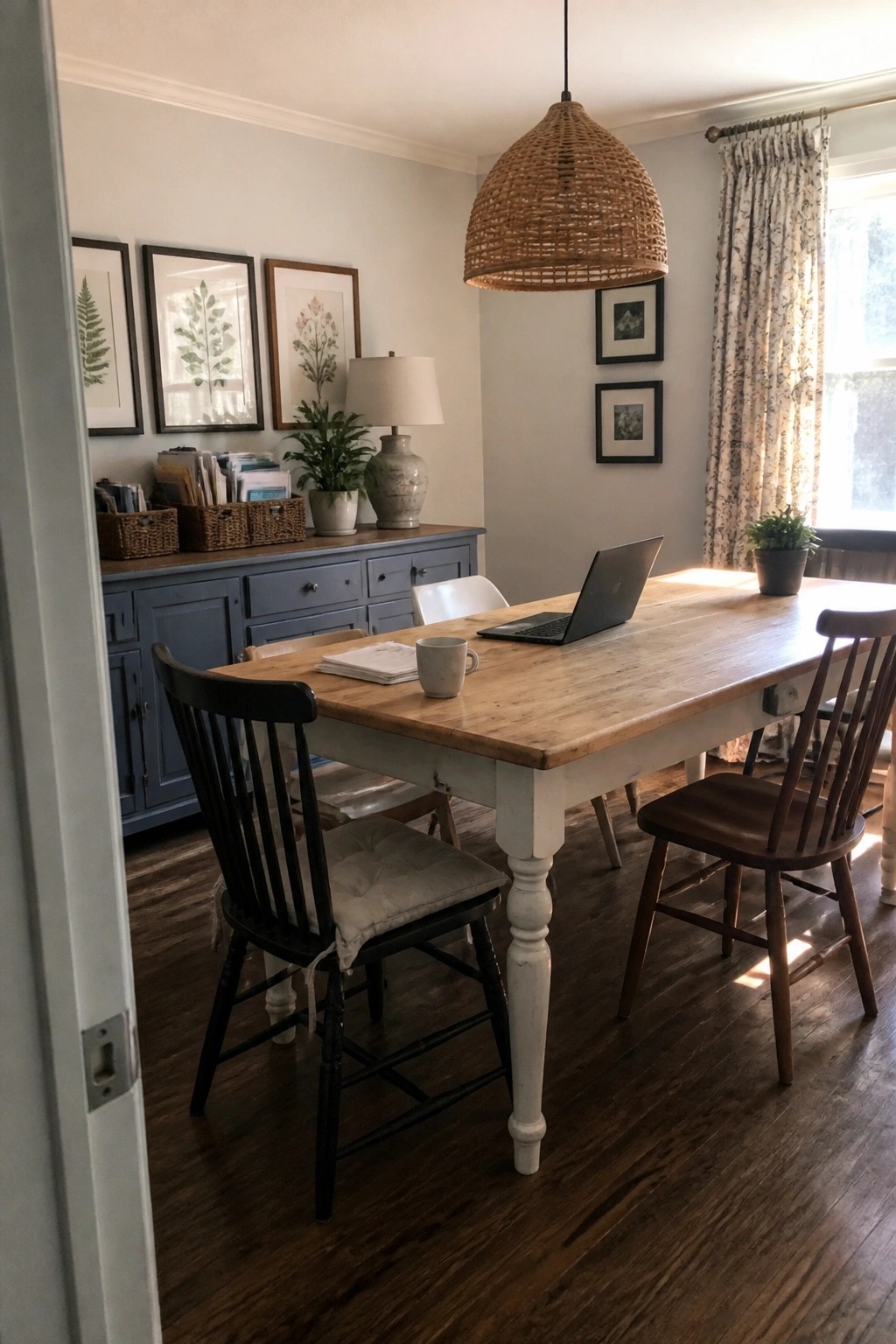 A light wood dining table with a laptop, mug, and papers sits in a room with mixed wooden chairs, a blue sideboard, and framed art on the walls.