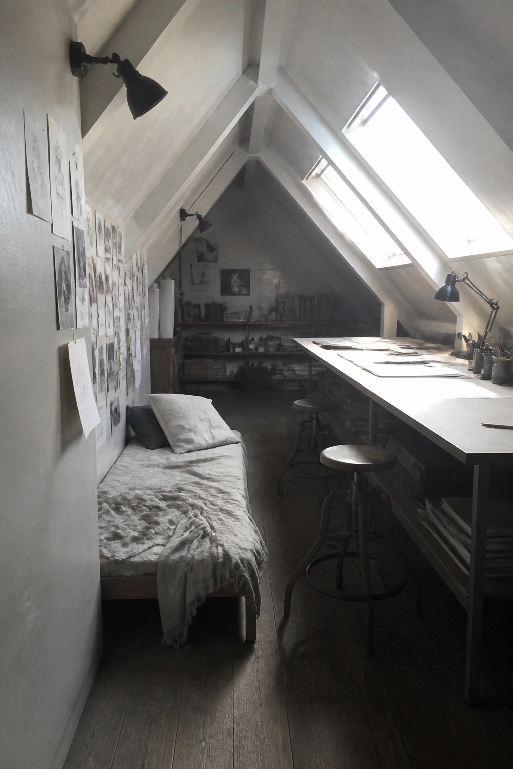An attic room with a long white work table along one wall, two wooden stools, a simple daybed, and several skylights in the sloped ceiling.