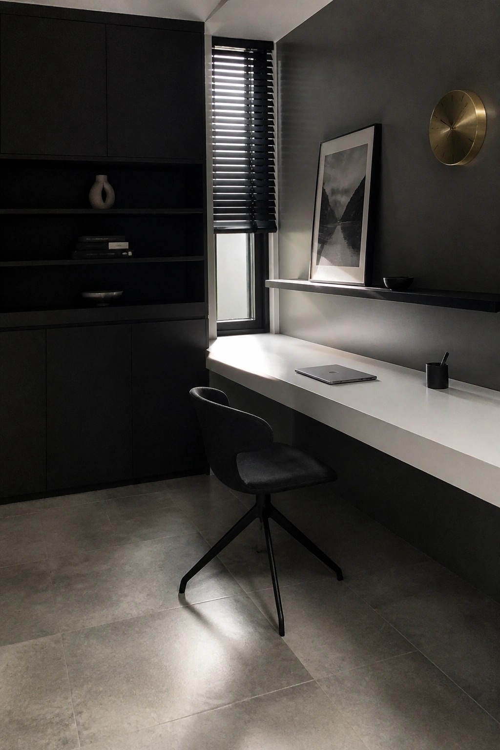 A modern home office with dark built-in cabinetry, open shelves, a white floating desk, a black swivel chair, and a window with blinds.