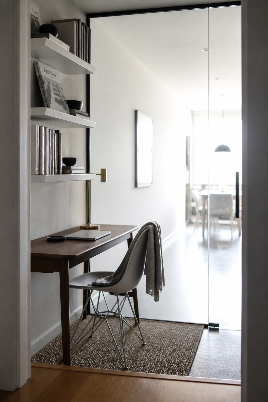 A compact wooden desk with white floating shelves above it holding books and small objects, a gray molded chair, and a woven rug on the floor near glass doors.