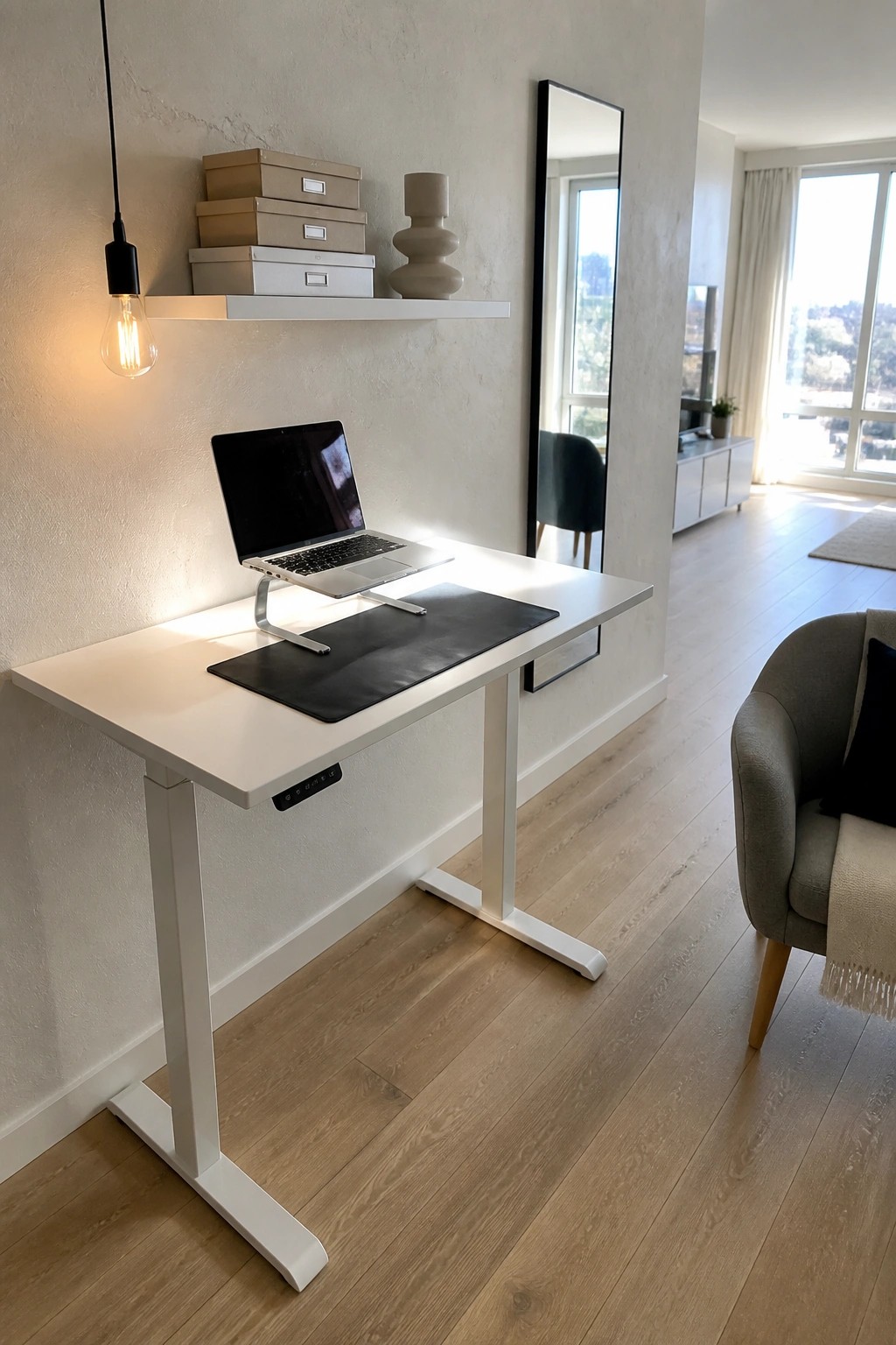 A white height-adjustable desk with an open laptop on a riser and a black desk mat, positioned under a floating shelf holding stacked storage boxes in a bright room.