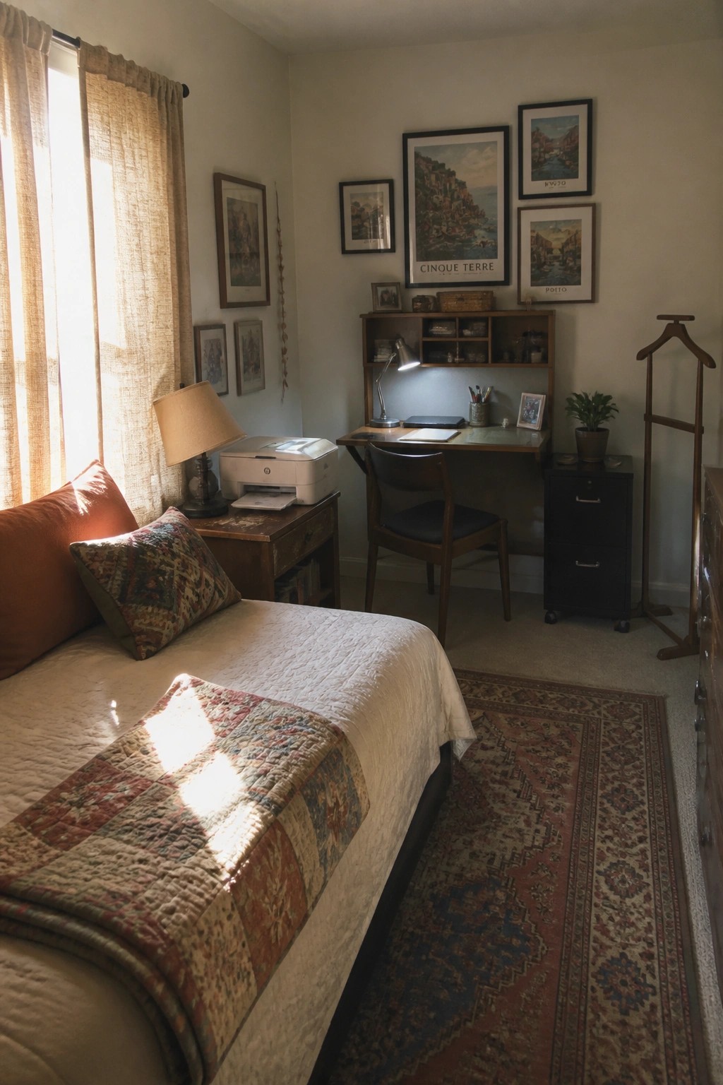 A bedroom interior showing a bed with a quilt on the left and a wooden desk with chair, shelves, and a printer in the right corner, with framed prints on the wall above the desk.