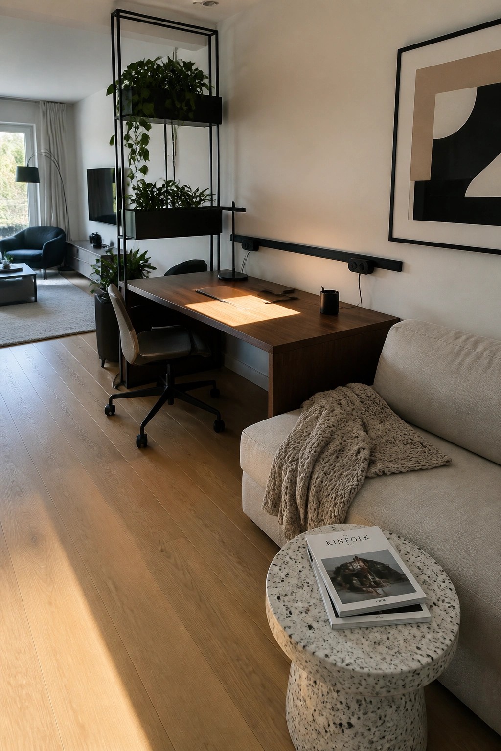 A wooden desk with an office chair is positioned next to a beige sofa in an open living room with wood floors and a tall plant shelf divider.