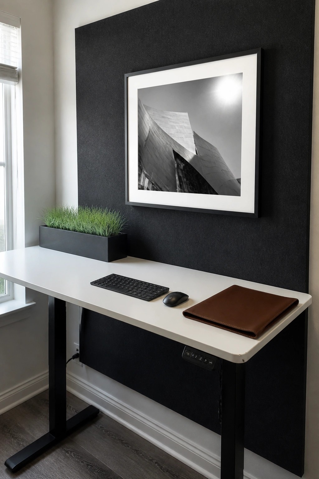 A white desk with a wireless keyboard, mouse, brown laptop sleeve, and black planter with grass sits against a black wall with a large illuminated black and white framed photograph above it.