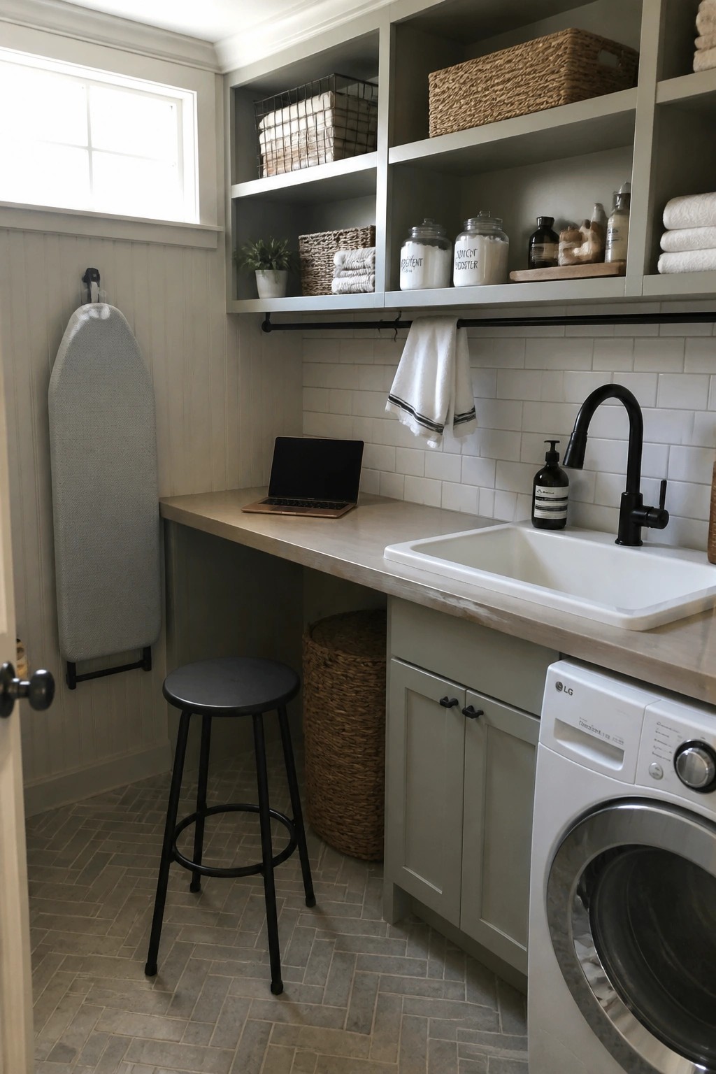 A laundry room interior with gray cabinets, a white sink, a washing machine, open shelves holding baskets and jars, a laptop on the counter, a black stool, and an ironing board mounted on the wall.