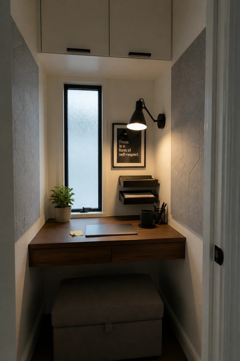 A narrow interior nook with a floating wooden desk, closed laptop, wall-mounted black lamp, potted plant on the left, and gray upholstered stool underneath.
