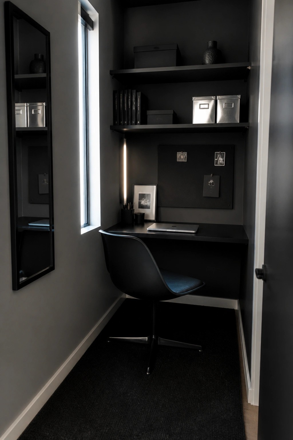 A narrow dark home office nook with built-in black shelves above a desk, a black swivel chair, and a tall vertical window on the left wall.
